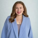 Woman with long wavy brown hair in blazer against light background.
