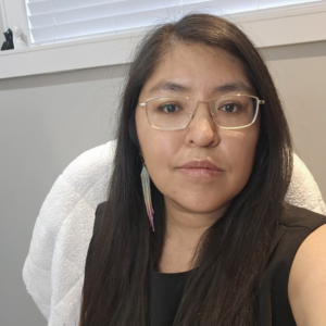 An Indigenous woman with glasses and long dark hair, wearing professional attire and smiling