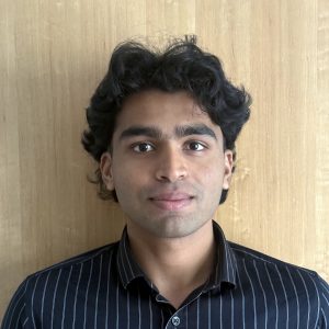 A South Asian man with thick, wavy black hair and a slight smile, wearing a dark pinstriped button-up shirt in front of a light wood-panelled wall. 